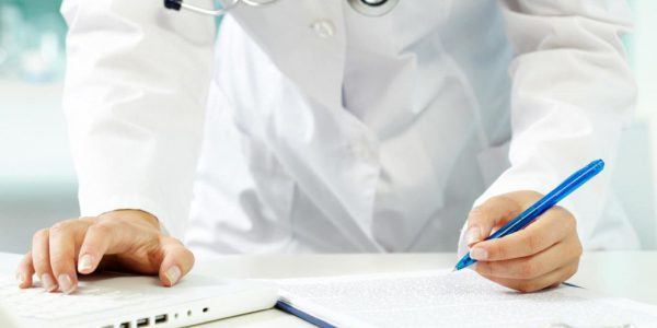 Médica olhando no computador e anotando as informações e em uma prancheta